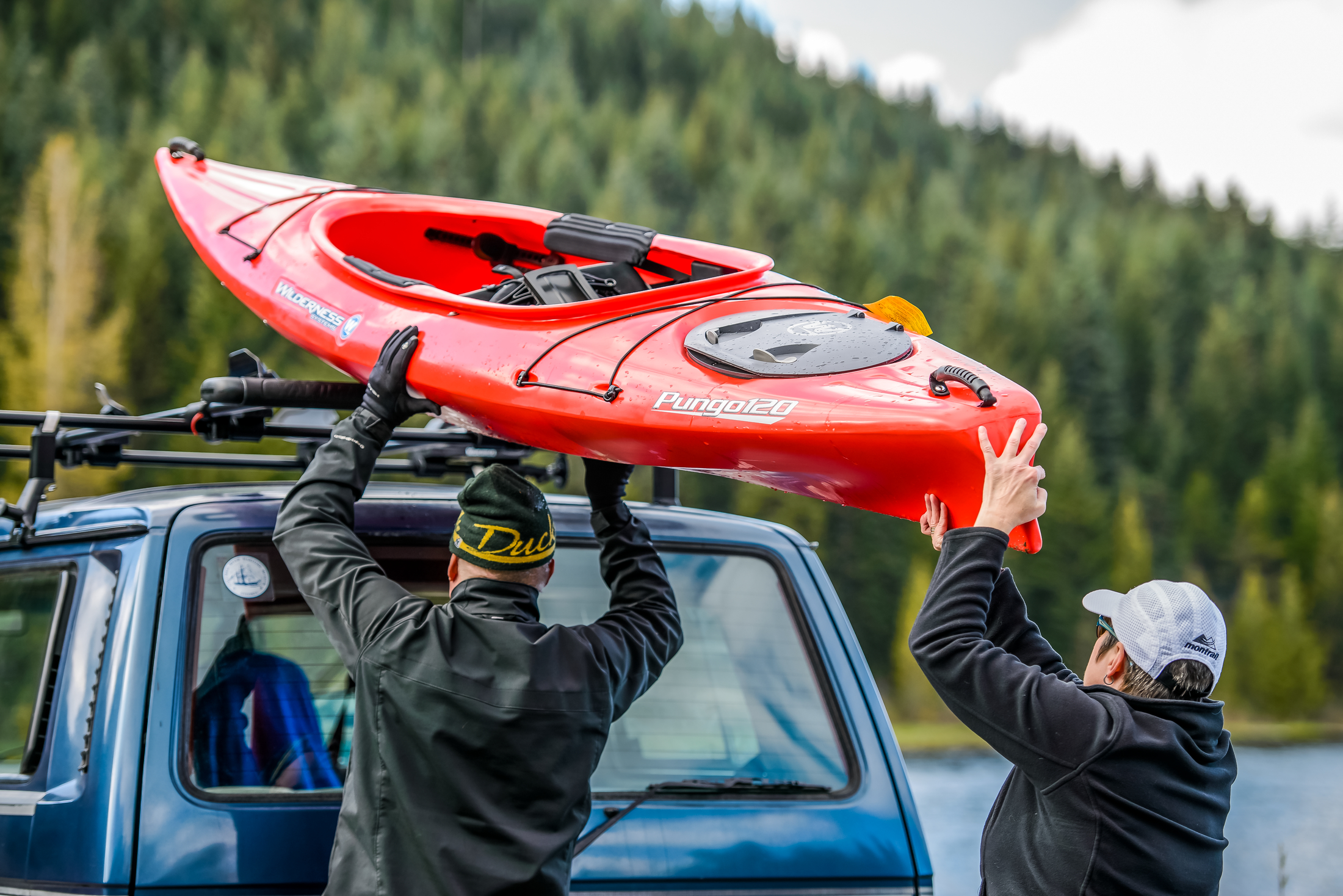 Kayak being loaded onto a vehicle roof rack beside a forested lake